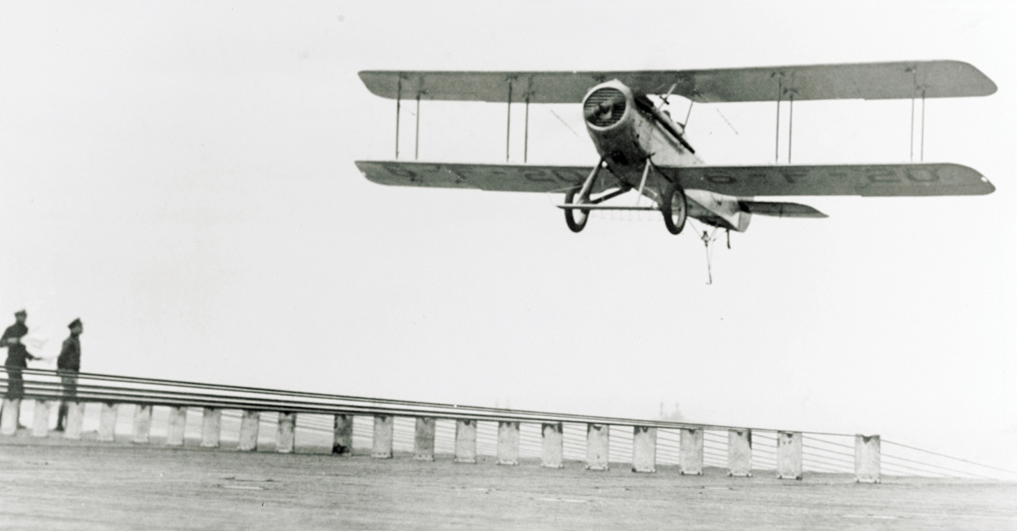 A VE-7 aircraft, using a tailhook, lands on USS Langley in May 1927, using longitudinal wires on fiddle bridges for an arresting arrangement.